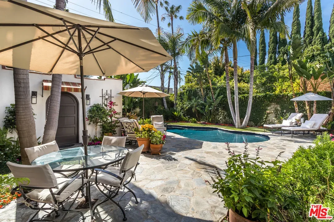 36 A backyard features a kidney-shaped pool, lounge chairs, umbrellas, and patio tables with chairs, surrounded by lush greenery and tall palm trees. A white stucco wall with a wooden door is in the background.